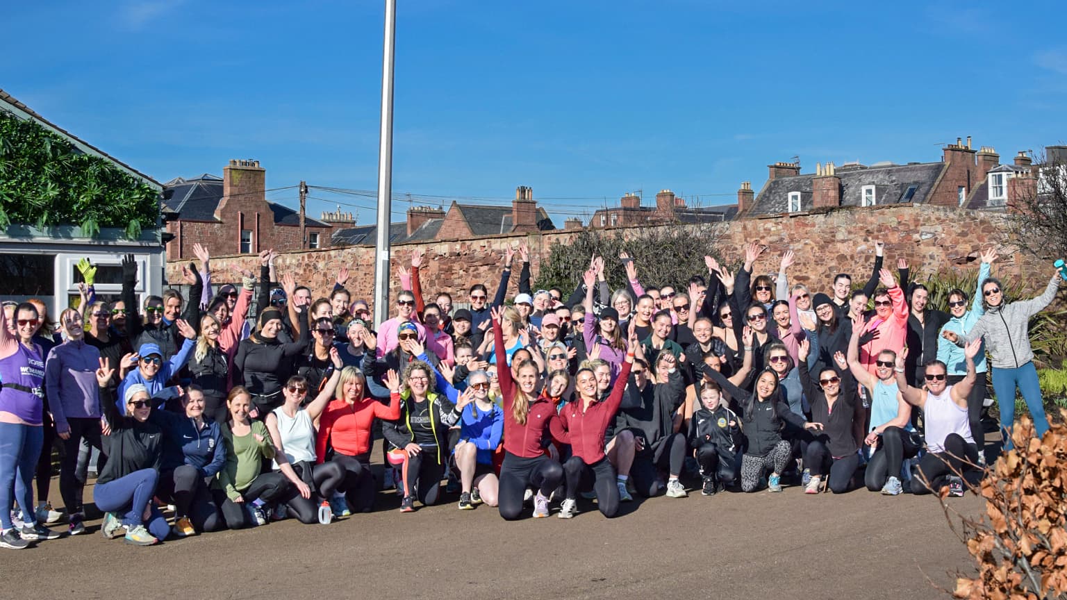 A large, diverse group of women in colourful running gear posing together and waving under a clear blue sky in East Lothian, capturing the community spirit of Run for Her.