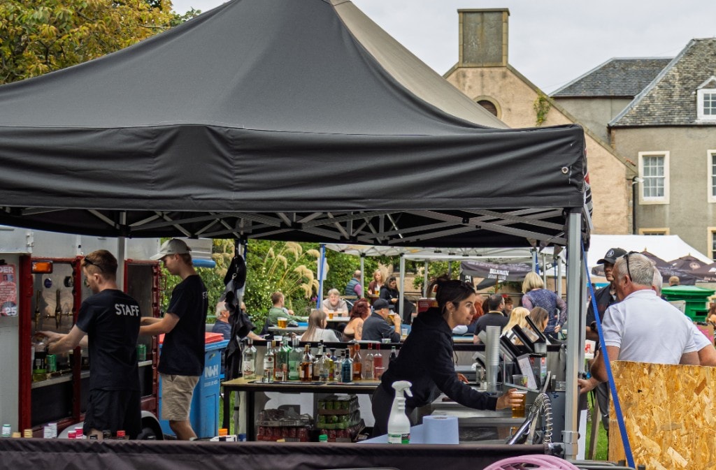Bar staff serving drinks beneath the black EH32 Live canopy, with Cockenzie House visible beyond the gardens.