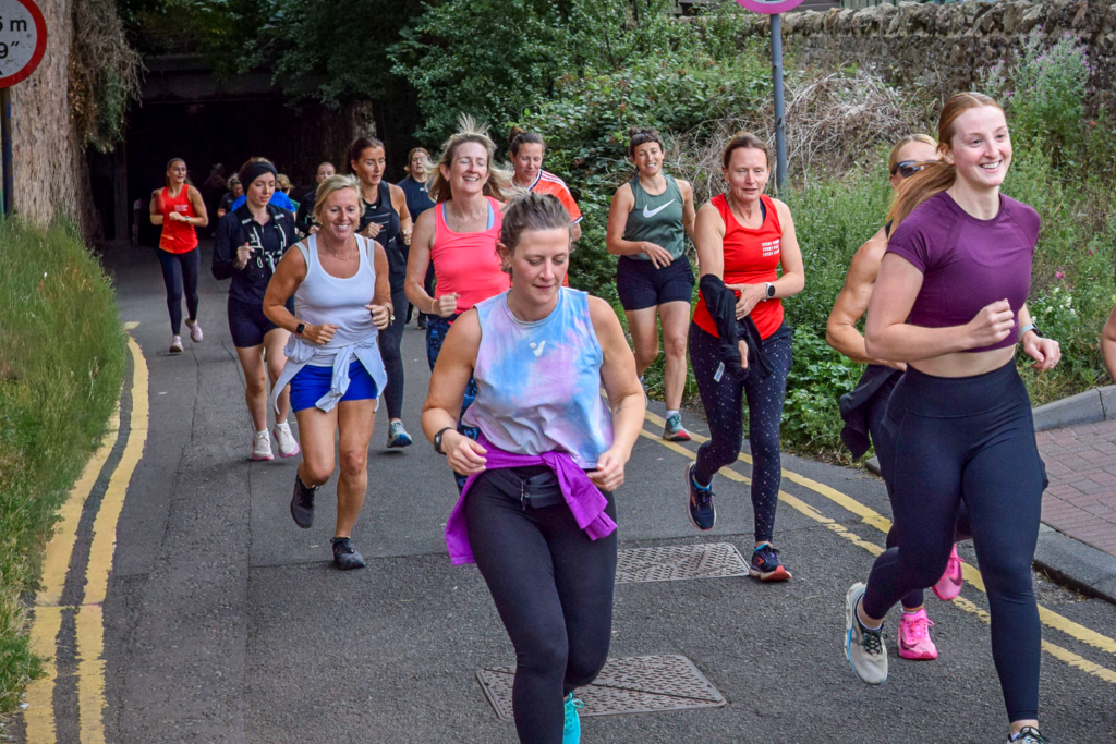 A large, diverse group of women jogging together on a paved path in East Lothian, smiling and talking as part of a Run for Her community event.